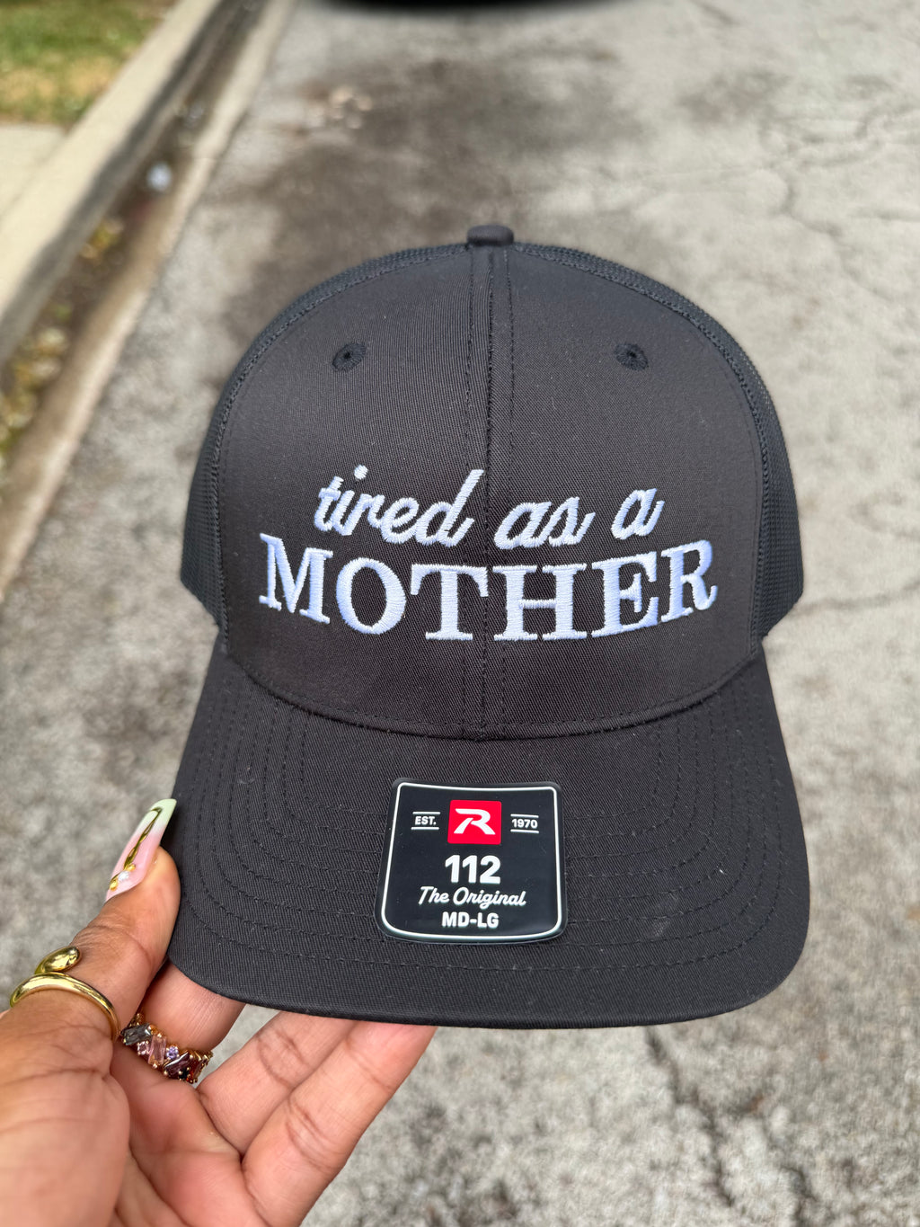 Black snapback cap with 'tired as a mother' text in light blue.